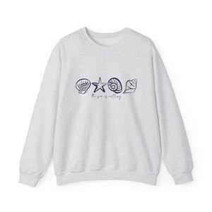 May include: A light gray sweatshirt with a navy blue graphic of seashells and a starfish. The text "the sea is calling" is printed below the graphic.