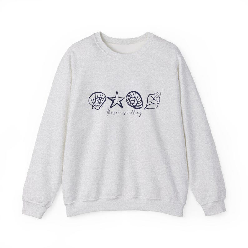 May include: A light gray sweatshirt with a navy blue graphic of seashells and a starfish. The text "the sea is calling" is printed below the graphic.