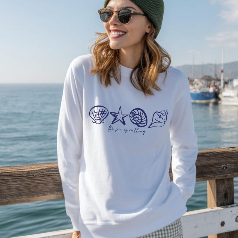 May include: White long-sleeve sweatshirt with a blue graphic of seashells, a starfish, and the text "the sea is calling".