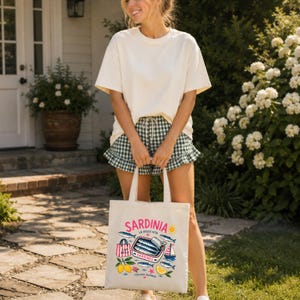May include: Cream-colored tote bag with the word "SARDINIA" in pink, featuring sardines, lemons, and a sailboat illustration. The bag is held by a person wearing a cream-colored t-shirt and green and white checkered shorts.