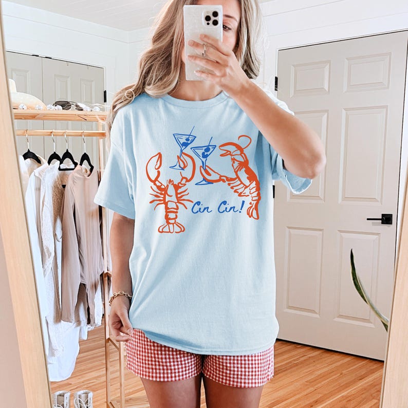 K&ouml;nnte beinhalten: Hellblaues T-Shirt mit einer Grafik von zwei orangefarbenen Hummern, die mit blauen Martinigl&auml;sern ansto&szlig;en. Der Text "Cin Cin!" ist darunter gedruckt. Das T-Shirt hat eine lockere Passform.