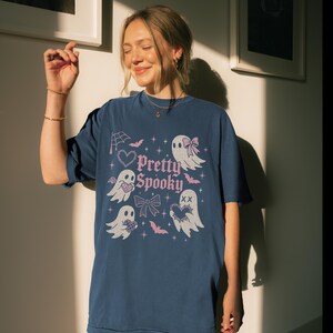 May include: Navy blue t-shirt with a "Pretty Spooky" design. The graphic features pink ghosts, bats, bows, and hearts. The shirt is a comfortable, casual style.