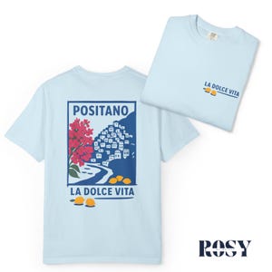 May include: Light blue t-shirts with a graphic design. One shirt displays a Positano illustration with red flowers, a coastal town, and lemons. The other shirt has "LA DOLCE VITA" text and lemons. The shirts are made of soft material.