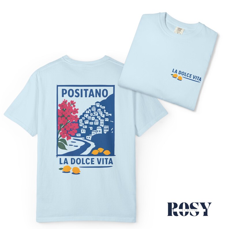 May include: Light blue t-shirts with a graphic design. One shirt displays a Positano illustration with red flowers, a coastal town, and lemons. The other shirt has "LA DOLCE VITA" text and lemons. The shirts are made of soft material.