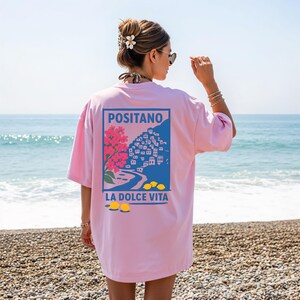 May include: A light pink t-shirt with a graphic of Positano, Italy, featuring a pink floral element, blue buildings, and the text "LA DOLCE VITA." The design is framed in a blue rectangle.