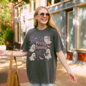 May include: Dark grey t-shirt featuring the text "Pretty Spooky" and pink and white illustrations of ghosts, bats, and bows. The shirt has a vintage, worn appearance.