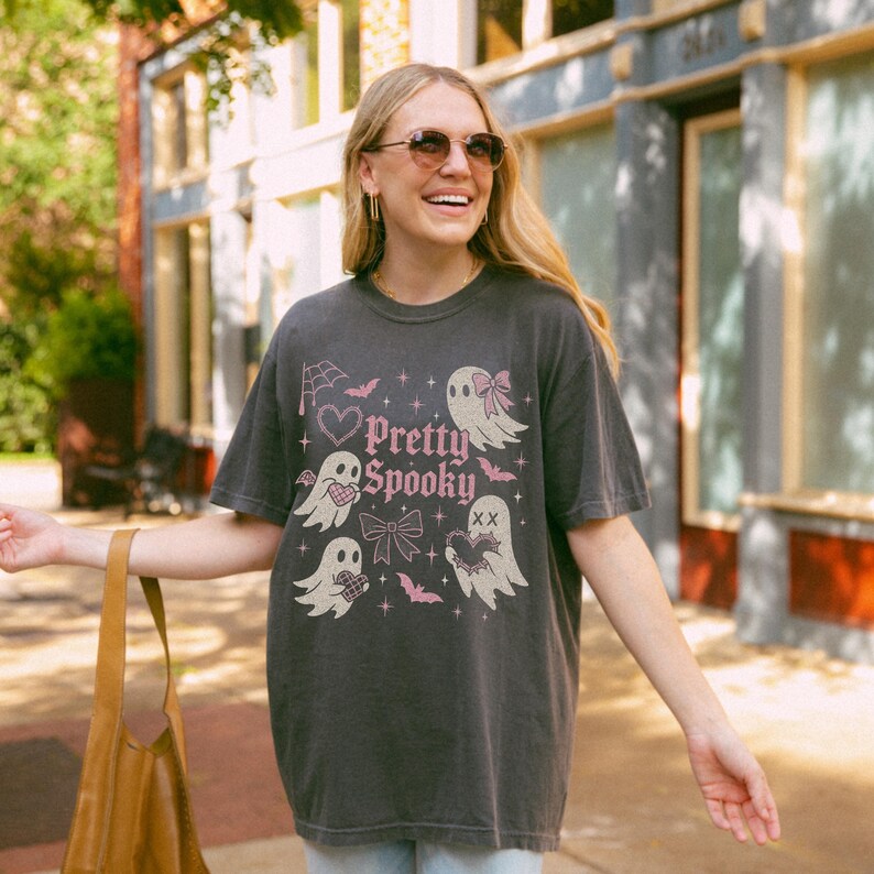 May include: Dark grey t-shirt featuring the text "Pretty Spooky" and pink and white illustrations of ghosts, bats, and bows. The shirt has a vintage, worn appearance.