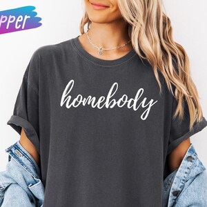 May include: Dark gray t-shirt with the word "homebody" in white script. The shirt has rolled-up sleeves and is worn with a light blue denim jacket. The word "pepper" is in a pink and blue box in the upper left corner.
