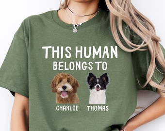 May include: Olive green t-shirt with the text "THIS HUMAN BELONGS TO" above two dog portraits. The dogs are a golden doodle and a Papillon. The names "CHARLIE" and "THOMAS" are printed below the dog portraits.