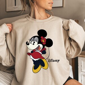 May include: A beige sweatshirt with a cartoon image of Minnie Mouse wearing a red dress and a black bow. The text "DISNEY" is printed below the image.