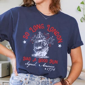 May include: Navy blue t-shirt with a graphic of a tall ship and the text "So Long, London. Had a Good Run. Signed, America *1776". The design includes red and white accents and stars.