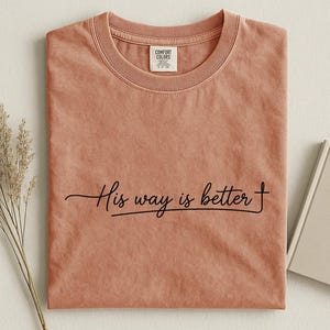 May include: A terracotta-colored short-sleeve t-shirt with a crew neckline. The shirt features the black cursive text "His way is better" printed across the center chest, with the final letter "r" extending into a cross symbol.