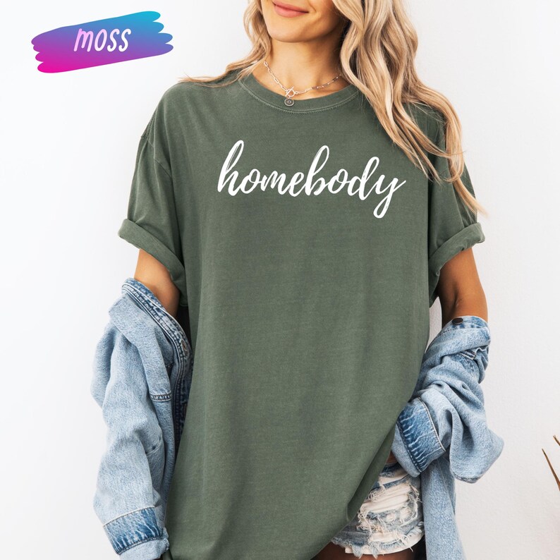 May include: Olive green t-shirt with the word "homebody" in white script. The shirt has rolled-up sleeves and is paired with a light blue denim jacket. A colorful "moss" logo is in the upper left corner.