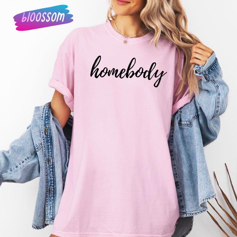 May include: A light pink t-shirt with the word "homebody" in black script. The shirt is worn with a denim jacket. The brand name "bloossom" is in the upper left corner in a blue and pink design.