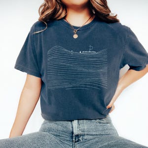 May include: A navy blue t-shirt featuring a minimalist white line drawing of a sailboat on wavy lines. The shirt has short sleeves and a relaxed fit. The design evokes a nautical theme.