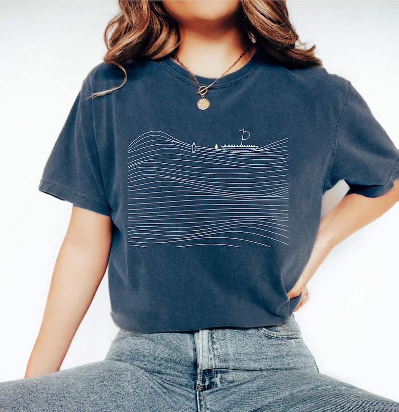 May include: A navy blue t-shirt featuring a minimalist white line drawing of a sailboat on wavy lines. The shirt has short sleeves and a relaxed fit. The design evokes a nautical theme.