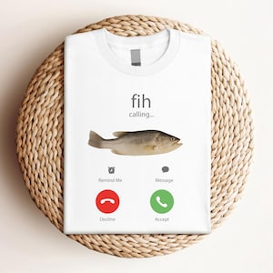 May include: White t-shirt with a humorous graphic of a fish, featuring the text "fih calling..." and a fish illustration. Below are icons for "Remind Me" and "Message", with "Decline" and "Accept" call buttons.