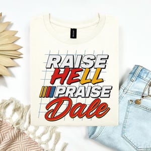 May include: Cream-colored t-shirt with the text "RAISE HELL PRAISE Dale" in a retro racing-style font. The design features red, yellow, and blue accents, and a grid background. The shirt is folded on a white surface.