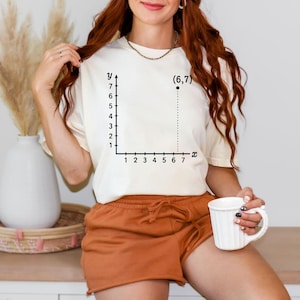 May include: Cream-colored t-shirt featuring a black graph with the coordinates (6,7). The graph has an x and y axis with numbers. The shirt is worn with orange shorts.