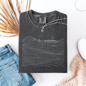 May include: A charcoal gray t-shirt with a white line drawing of a boat on wavy lines. A silver necklace with a circular pendant rests on the shirt. The shirt is folded next to blue denim shorts and white sneakers.
