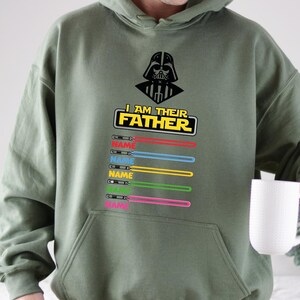 Pu&ograve; includere: Felpa con cappuccio verde oliva con grafica nera di Darth Vader e la scritta "I AM THEIR FATHER". Sotto il testo ci sono disegni di spade laser colorate con la parola "NAME" stampata su ciascuna. La felpa ha una tasca frontale.