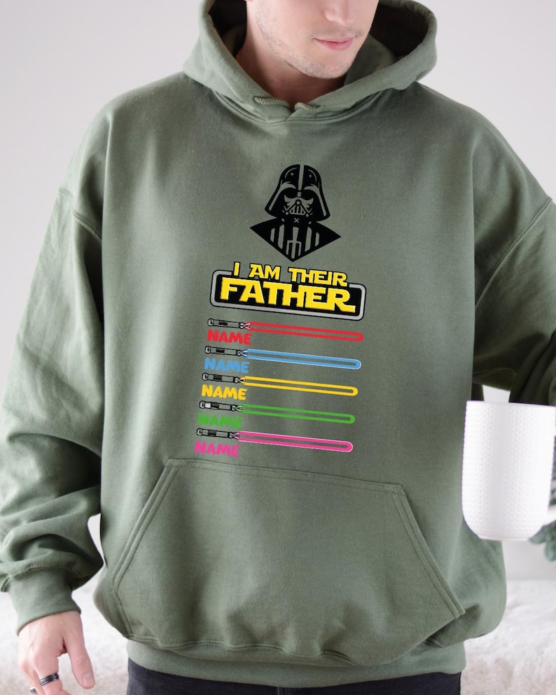 Pu&ograve; includere: Felpa con cappuccio verde oliva con grafica nera di Darth Vader e la scritta "I AM THEIR FATHER". Sotto il testo ci sono disegni di spade laser colorate con la parola "NAME" stampata su ciascuna. La felpa ha una tasca frontale.