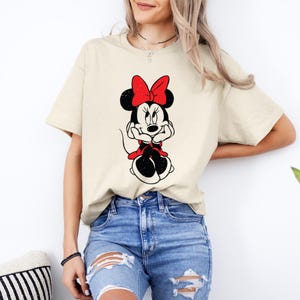 May include: A beige t-shirt with a black and white cartoon image of Minnie Mouse. Minnie Mouse is wearing a red bow and has a black and white polka dot dress.