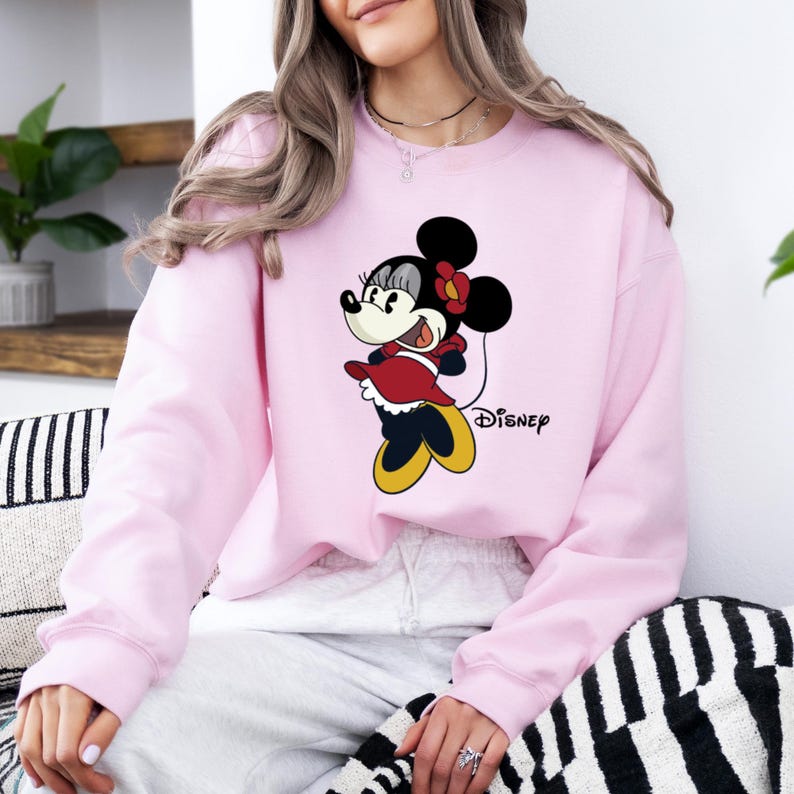May include: Pink sweatshirt with a cartoon image of Minnie Mouse in a red dress and yellow shoes. The text "DISNEY" is printed on the bottom right corner of the sweatshirt.