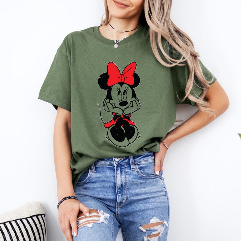 May include: A green t-shirt with a cartoon image of Minnie Mouse wearing a red bow. Minnie Mouse is sitting with her hands on her chin and has a black and white color scheme.