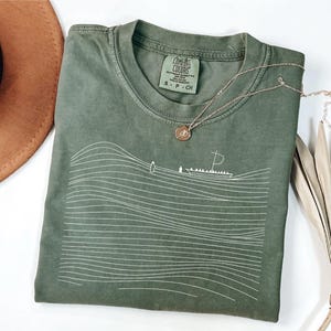 May include: Olive green t-shirt with a white line drawing of waves and a boat. The shirt has a crew neck and a small tag on the collar. A gold necklace with a pendant is draped across the shirt.