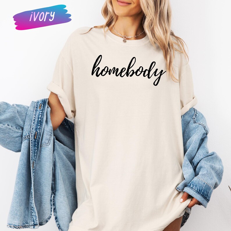 May include: Ivory-colored t-shirt with the word "homebody" in black script. The shirt is a relaxed fit. A denim jacket is draped over the arm. The word "ivory" is written in a colorful banner in the upper left corner.