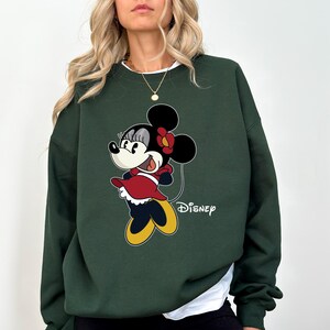 May include: A dark green sweatshirt with a cartoon image of Minnie Mouse wearing a red dress and a yellow bow. The text "DISNEY" is printed below the image.