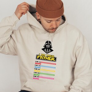 Pu&ograve; includere: Felpa con cappuccio beige con grafica nera di Darth Vader e la scritta "I AM THEIR FATHER". Sotto, disegni di spade laser colorate con la parola "NAME" stampata su ciascuna. La felpa ha un cappuccio con coulisse e una tasca frontale.