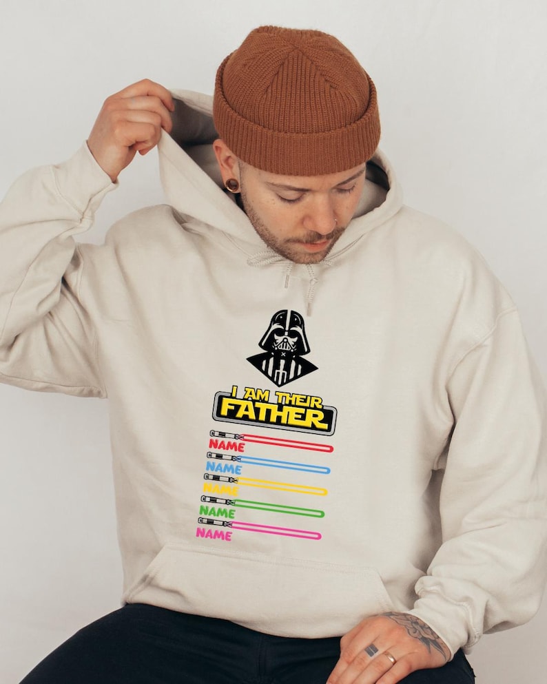 Pu&ograve; includere: Felpa con cappuccio beige con grafica nera di Darth Vader e la scritta "I AM THEIR FATHER". Sotto, disegni di spade laser colorate con la parola "NAME" stampata su ciascuna. La felpa ha un cappuccio con coulisse e una tasca frontale.