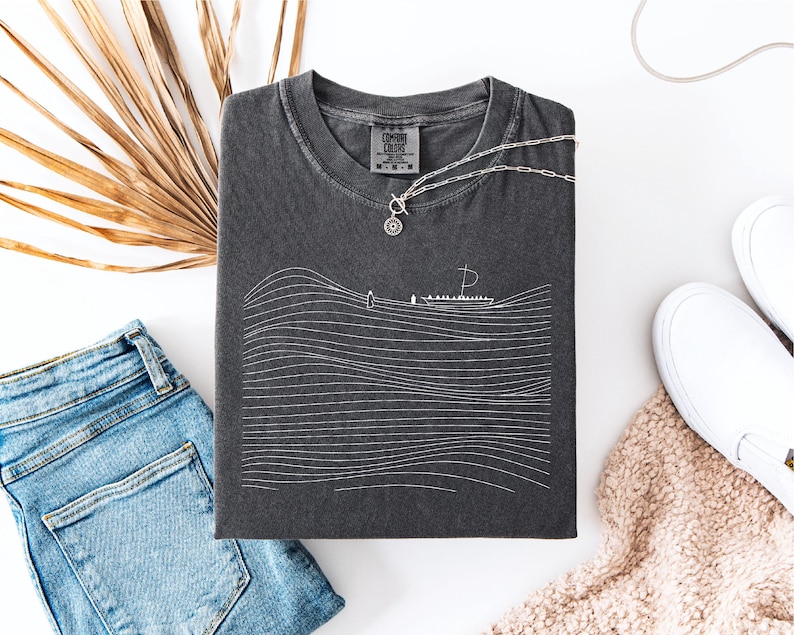May include: Dark gray t-shirt with a white line drawing of a boat on wavy lines. A silver necklace with a circular pendant rests on the shirt. The shirt is folded next to blue denim shorts and white sneakers.