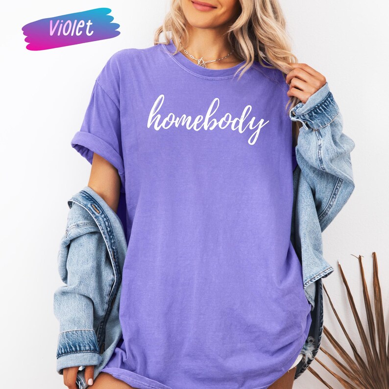 May include: A violet-colored t-shirt with the word "homebody" in white script. The shirt has rolled-up sleeves and is paired with a denim jacket. A colorful "Violet" banner is in the upper left corner.