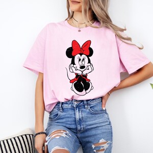 May include: A pink t-shirt with a black and white image of Minnie Mouse wearing a red bow. Minnie Mouse is sitting with her hands on her chin.