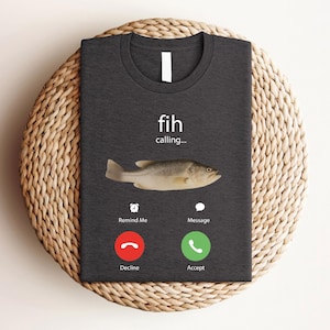 May include: Dark gray t-shirt featuring a fish graphic with the text "fih calling...". The design includes a fish image and a phone interface with "Decline" and "Accept" buttons. The shirt is displayed on a woven, circular mat.