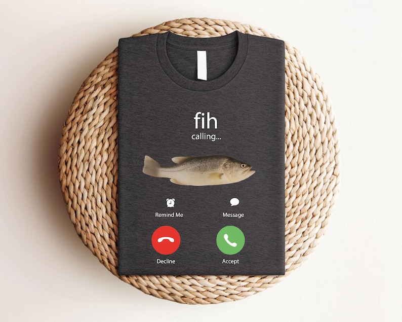 May include: Dark gray t-shirt featuring a fish graphic with the text "fih calling...". The design includes a fish image and a phone interface with "Decline" and "Accept" buttons. The shirt is displayed on a woven, circular mat.
