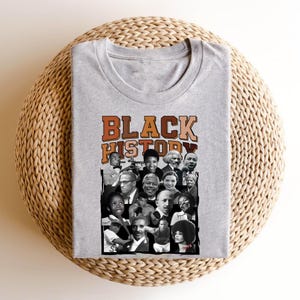 May include: A gray t-shirt with a collage of black and white photos of prominent African Americans. The text "BLACK HISTORY" is printed in orange, brown, and black letters across the top of the collage.