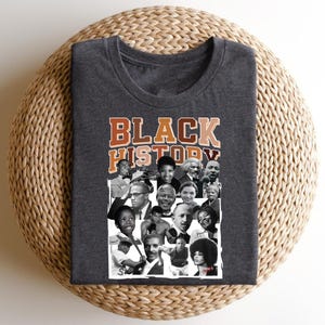 May include: A dark gray t-shirt with the text "BLACK HISTORY" in orange and brown letters. The shirt features a collage of black and white photos of notable African Americans.