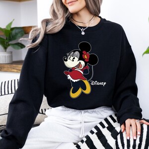 May include: Black sweatshirt with a cartoon image of Minnie Mouse in a red dress and a yellow bow. The text "DISNEY" is printed on the bottom right corner of the sweatshirt.