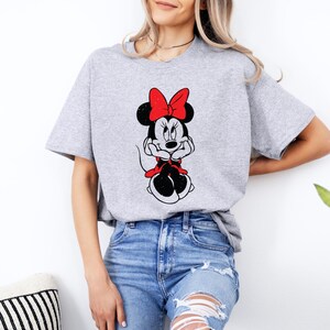 May include: A gray t-shirt with a black and white image of Minnie Mouse wearing a red bow. Minnie Mouse is sitting with her hands clasped in front of her.
