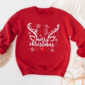 May include: Red crewneck sweatshirt with a white reindeer design. The design includes antlers, snowflakes, a red nose, and the text "Trump Merry Christmas". A festive holiday-themed apparel item.