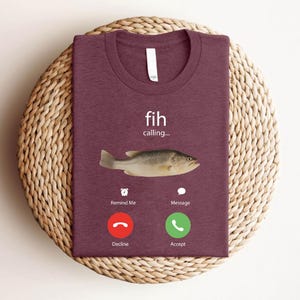 May include: A maroon t-shirt with a crew neck, featuring a fish graphic and the words "fih calling...". Below the fish are two phone-style buttons labeled "Decline" and "Accept". The shirt is folded on a woven, circular placemat.