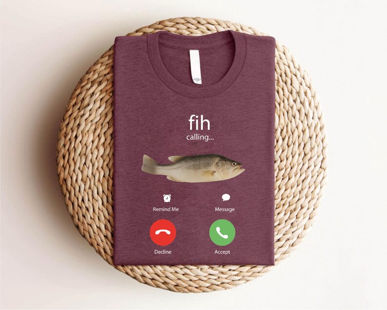 May include: A maroon t-shirt with a crew neck, featuring a fish graphic and the words "fih calling...". Below the fish are two phone-style buttons labeled "Decline" and "Accept". The shirt is folded on a woven, circular placemat.