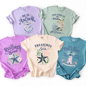 May include: Five short-sleeve crew neck t-shirts in lavender, light green, light purple, cream, and teal. Each shirt features a unique graphic print with religious text and coastal illustrations including an anchor, a rock, a starfish, and a lighthouse.