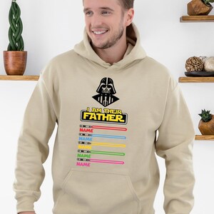 Pu&ograve; includere: Una felpa con cappuccio beige con una grafica nera di Darth Vader e la scritta "I AM THEIR FATHER". Sotto ci sono spade laser colorate con la parola "NAME" stampata su ciascuna. La felpa ha una tasca frontale e un cappuccio.
