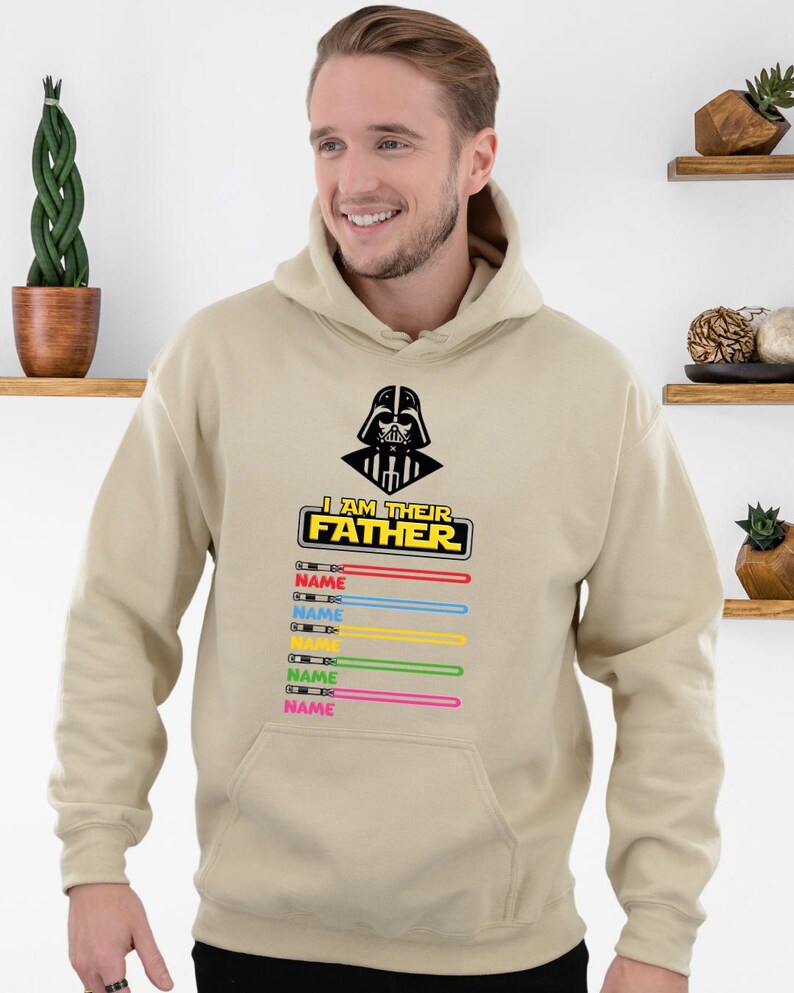 Pu&ograve; includere: Una felpa con cappuccio beige con una grafica nera di Darth Vader e la scritta "I AM THEIR FATHER". Sotto ci sono spade laser colorate con la parola "NAME" stampata su ciascuna. La felpa ha una tasca frontale e un cappuccio.