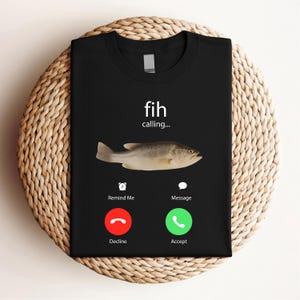 May include: Black t-shirt featuring a fish graphic with the text "fih calling...". Below the fish are icons for "Remind Me" and "Message", with "Decline" and "Accept" buttons. The shirt is displayed on a woven, circular surface.